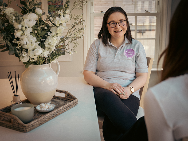 Sam Shenton - Sorted by Sam. Shoulder length brown hair, glasses, wearing black trousers and a grey polo top with the sorted! By Sam logo. She is sat next to a table with a tray containing a vase with white flowers, a candle and a reed diffuser